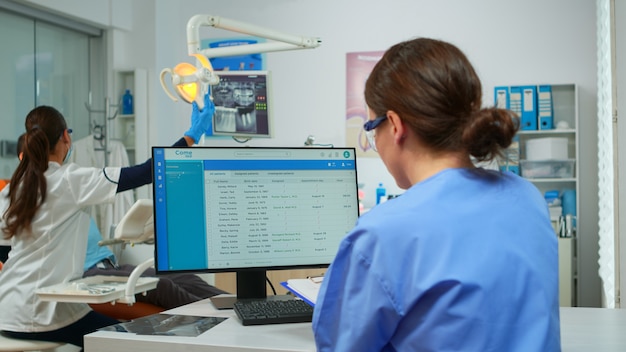 Ассистент ортодонта делает заметки в буфере обмена, проверяет назначения, в то время как врач-стоматолог с маской для лица осматривает пациента с зубной болью, сидящего на стоматологическом кресле.