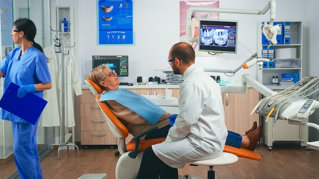 Orthodontist discussing MRI scan with patient sitting in stomatological chair in dental clinic. Elderly woman explaining dental problem to doctor indicating mouth while nurse working in background