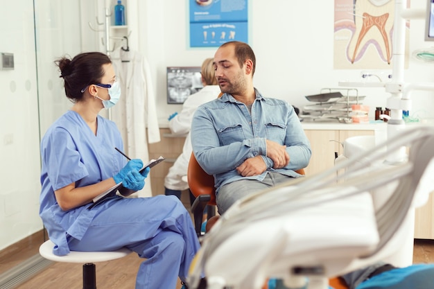 Медсестра-ортодонт обсуждает с пациентом-мужчиной о консультации с зубом, записывая лечение в буфер обмена, сидя в стоматологическом кабинете