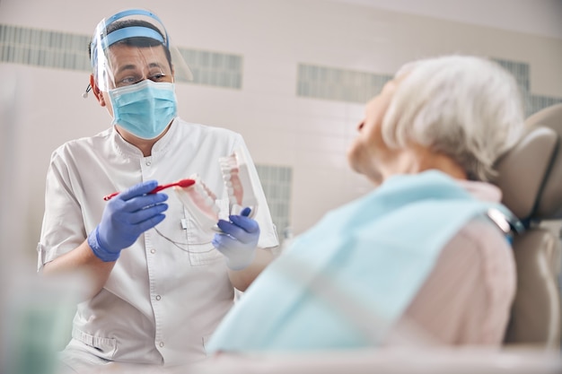 Ortodontista che indossa l'uniforme e tiene in mano un modello di denti sovradimensionati e uno spazzolino da denti nel suo ufficio