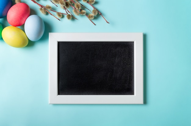 Orthodox Easter concept. Colorful eggs and pussy willow branches on blue background with empty chalkboard.