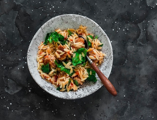 Orzo pasta with meatballs and spinach in tomato sauce on a dark background top view