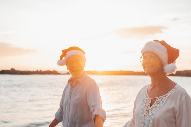 Oud schattig stel volwassen personen die genieten en plezier hebben samen op het strand met kerstmutsen op vakantiedagen Wandelen op het strand met de zonsondergang op de achtergrond bij winterxA