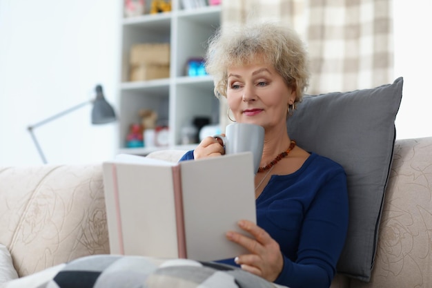 Oude dame die een boek leest en een kopje koffie drinkt terwijl ze thuis op de bank zit