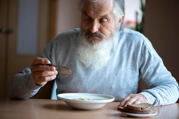 Foto oude droevige man met een lange grijze baard die bij de tafel zit en soep en brood eet