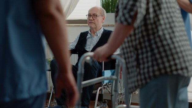 Foto oude gehandicapte man ontspannen in verpleeghuis kamer. gepensioneerde persoon met een handicap die in een rolstoel zit en zich voorbereidt op herstel en welzijn terwijl verpleegkundigen de patiënt helpen met een looprek