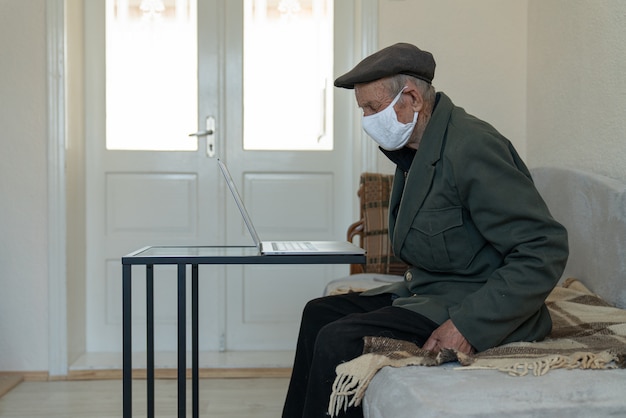 Oude gepensioneerde man in beschermende medische gezichtsmasker zittend op de bank thuis in de buurt van de werktafel met laptop