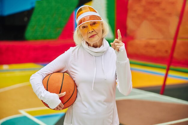 Oude, gezonde en vrolijke dame met basketbalbal op een kleurrijk plein buitenshuis