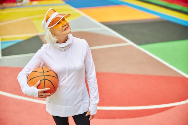 Oude, gezonde en vrolijke dame met basketbalbal op een kleurrijk plein buitenshuis