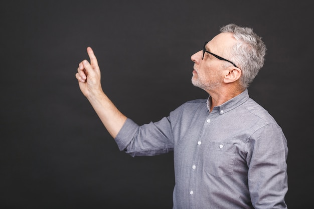 Oude hogere mens die glazen dragen die tegen zwarte verbaasde muur worden geïsoleerd en glimlachen terwijl het voorstellen met hand en met vinger richten.