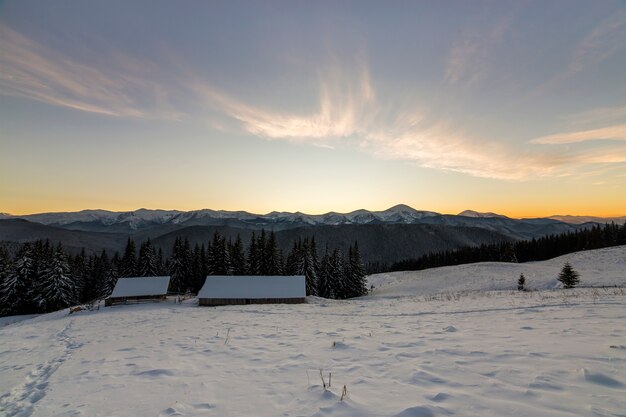 Oude houten huis, hut en schuur in diepe sneeuw op bergvallei, vuren bos, bosrijke heuvels op heldere blauwe hemel bij zonsopgang. Berglandschap winter panorama.