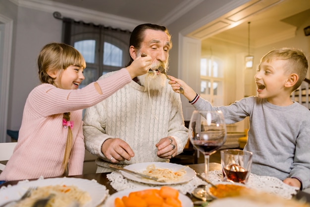 Oude knappe grootvader met zijn twee kleinkinderen zitten aan de keukentafel en eten pasta. Klein meisje en jongen grootvader voeden met pasta en lachen