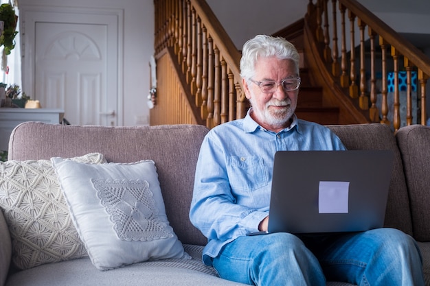 Oude man glimlachend zittend op de bank in de woonkamer met behulp van laptop, genietend van werken met de computer, tevreden voelen met het verzenden van berichten, vrienden bellen, surfen op het web online concept