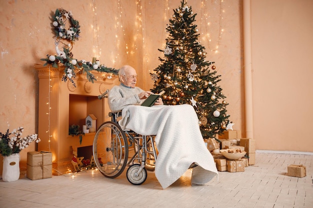 Oude man in een rolstoel die bij de kerstboom zit en een boek leest