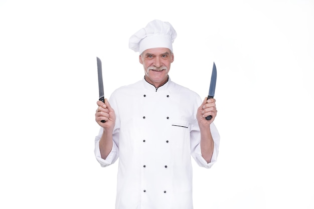 Oude man, professionele chef-kok in uniform die twee metalen mes vasthoudt terwijl hij op de witte muur blijft