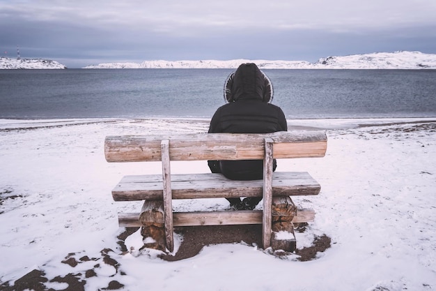 Oude man zit alleen op een bankje tegen de achtergrond van de baai. concept van eenzaamheid en eenzaamheid. Koud winterlandschap. Een man zit alleen en kijkt naar de Noordelijke IJszee