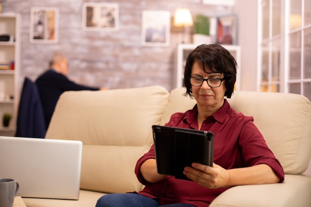 Oude oudere vrouw die op de bank zit en een digitale tablet-pc gebruikt in een gezellige woonkamer.