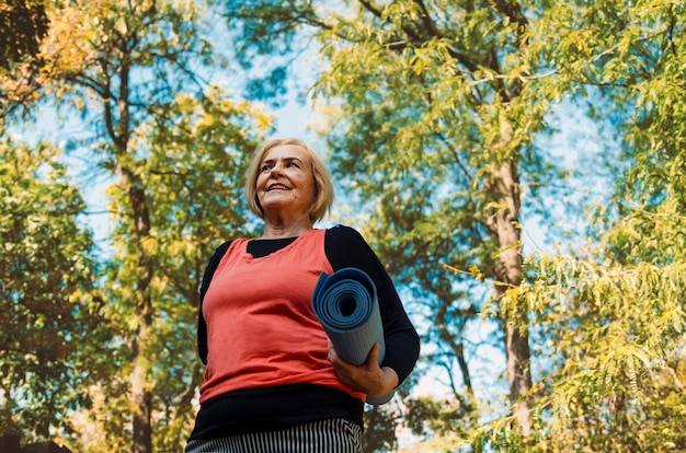 Oude senior vrouw met yoga mat buiten in het bos gaan op training. gezonde levensstijl en lichaamsbeweging bij pensionering. grijs haar gerijpt vrouwelijke training buitenshuis