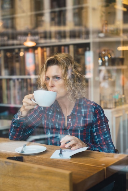 Oude volwassen vrouw die alleen koffie drinkt met een boek dat wegkijkt van de camera. schot genomen door een glas