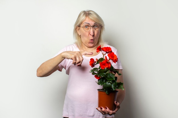 Oude vriendelijke vrouw in een casual T-shirt en een bril houdt kamer bloem en wijst de vinger op het met een verbaasd gezicht op een geïsoleerde lichte muur. Emotioneel gezicht. Concept van plantenverzorging, huis tuin