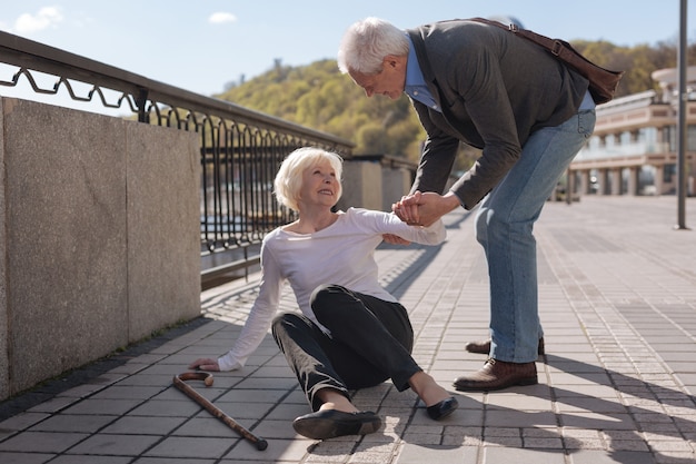 Oude vrij vrolijke vrouw die op de grond ligt en glimlacht terwijl beleefde echtgenoot haar helpt