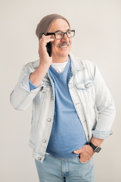 Oude vrolijke man in casual pullover, jasje, spijkerbroek en muts belt via smartphone terwijl hij bij een witte muur staat