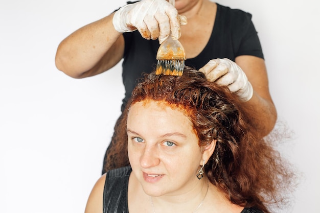 Oude vrouw die achter jonge vrouw staat en haar in het rood verven met borstel in de hand met latex witte handschoenen op witte achtergrond Haarkleuring thuis Het wegwerken van grijs haar Antiage