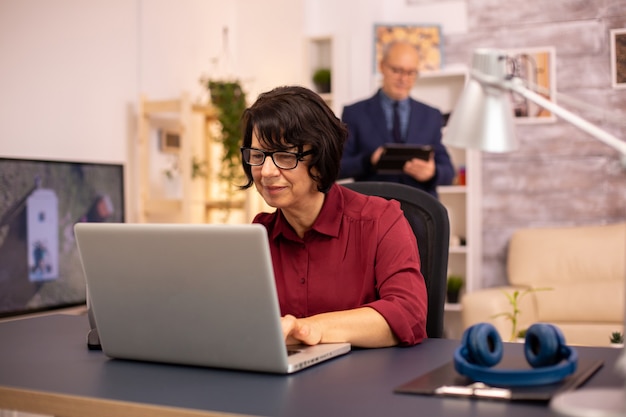 Oude vrouw die een moderne computer in haar woonkamer gebruikt terwijl haar man op de achtergrond loopt