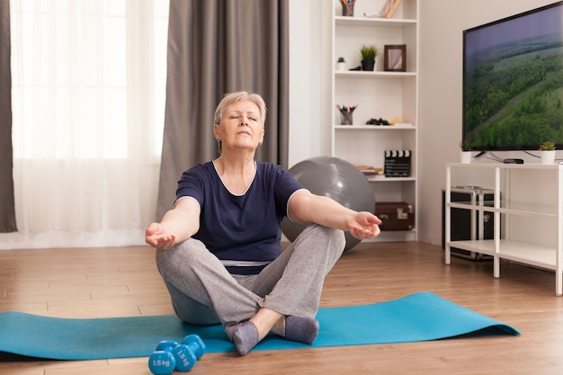 Oude vrouw die thuis op yogamat mediteert