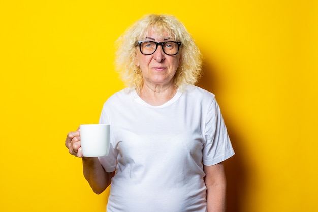 Oude vrouw in wit t-shirt en glazen houdt een kopje thee op een gele muur.