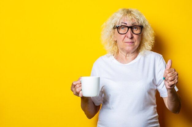 Oude vrouw in wit t-shirt en glazen houdt een kopje thee op een gele muur.