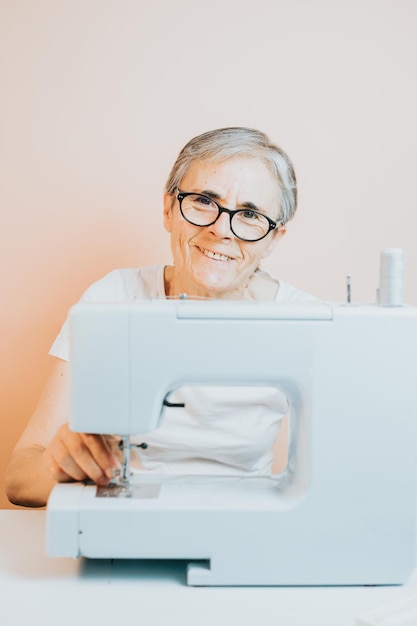 Oude vrouw kleermaker bezig met naaimachine glimlachend blij naar camera terwijl naaien Hergebruik oude stoffen voor nieuwe kleding duurzaamheid Oude mensen nieuwe hobby thuis senioren mentale activiteit en baan