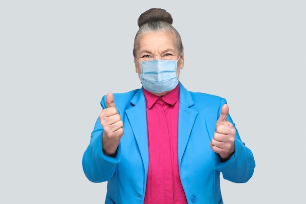 Oude vrouw met chirurgisch medisch masker dat als teken omhoog duimt. grootmoeder met lichtblauw pak en roze shirt, verzameld knotgrijs haar. indoor studio-opname, geïsoleerd op een grijze achtergrond