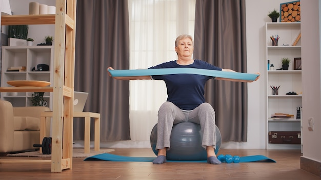 Oude vrouw oefenen met weerstand band zittend op fitness bal. oude vrouw tillen opleiding gezonde levensstijl sport fitness workout thuis met gewichten halter activiteit