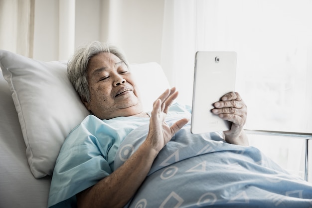Oude Vrouwelijke geduldige videovraag met tablet terwijl het liggen op bed in het ziekenhuis.
