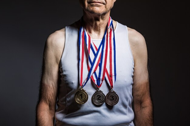 Oudere atleet met witte tanktop, met zonnevlekken op de armen, met drie medailles in de nek, die ze laten zien, op een donkere achtergrond. Sport- en overwinningsconcept