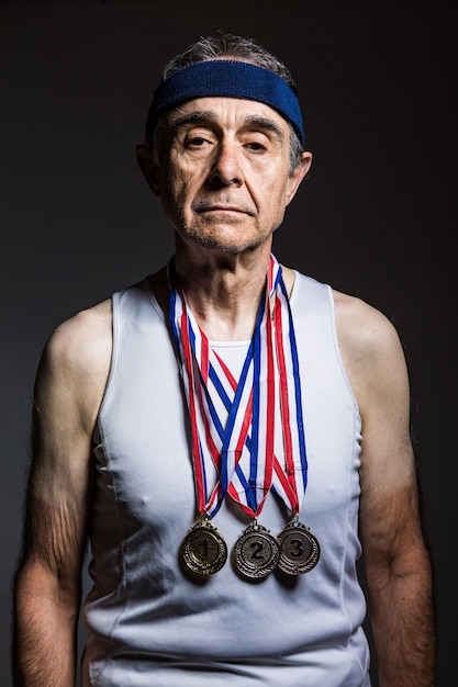 Oudere atleet met witte tanktop, met zonnevlekken op de armen, met drie medailles in de nek, die ze laten zien, op een donkere achtergrond. sport- en overwinningsconcept