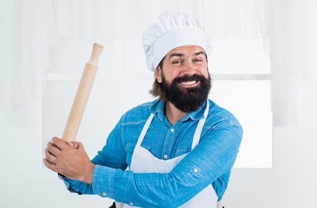 Oudere, bebaarde hipster draagt een koksmuts tijdens het koken van voedsel in de keuken met behulp van keukengerei met deegroller