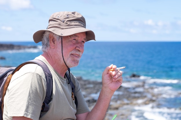 Oudere bebaarde man met hoed zittend op de rotsen bij de zee een sigaret rokend terwijl hij uitrust na een trekking langs de kliffen