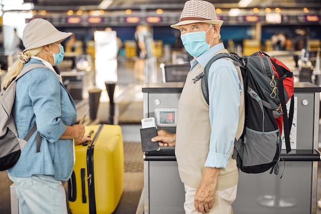 Oudere blanke man met gezichtsmasker die wacht tot zijn vrouw hun bagage aflevert