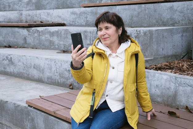 Oudere brunette vrouw zit in een stadspark op de trap glimlachend terwijl ze op een videolink praat