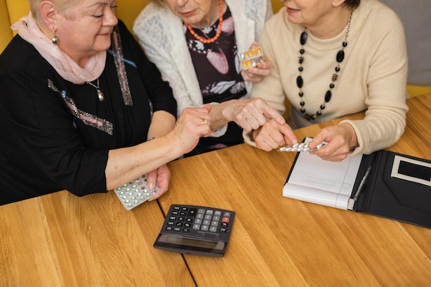 Oudere donkere en blonde vrouwen met pillen in hun handen en rekenmachine op tafel, bespreken medicijnen, prijzen. medische problemen en gezondheid terwijl ze hun ontmoeting vieren in een modern en gezellig café