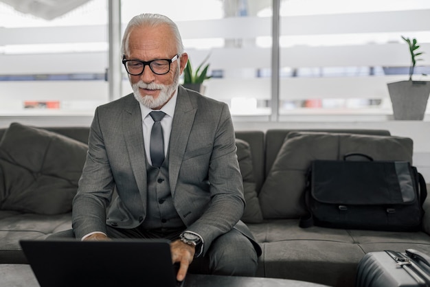 Oudere glimlachende zakenman die op laptop werkt terwijl hij op de bank op kantoor zit