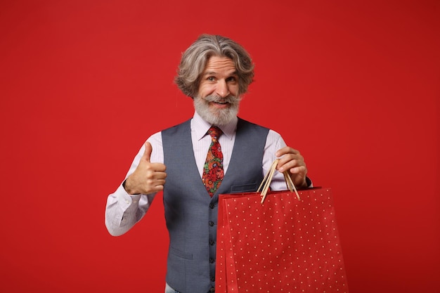 Oudere grijsharige snor bebaarde man in overhemd vest stropdas geïsoleerd op rode achtergrond. Mensen levensstijl concept. Bespotten kopie ruimte. Houd de pakkettas vast bij de aankoop na het winkelen en laat de duim omhoog zien.