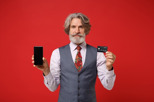 Oudere grijsharige snor bebaarde man in overhemd vest stropdas poseren geïsoleerd op rode achtergrond. Mensen levensstijl concept. Bespotten kopie ruimte. Houd mobiele telefoon vast met leeg leeg scherm, creditcard.