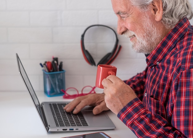 Oudere man die door sociale media-inhoud bladert met behulp van een laptop op een wit bureau dat een koffiekopje drinkt