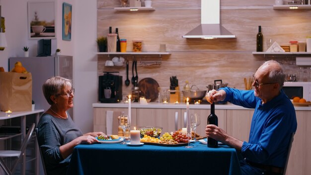 Oudere man die een fles rode wijn opent tijdens een romantisch diner. senior oud stel praten, aan tafel zitten in de keuken, genieten van de maaltijd, hun jubileum thuis vieren met gezond voedsel.