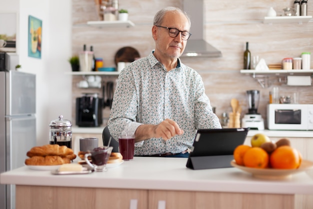 Oudere man die lacht met behulp van tablet die geniet van moderne internet online technologie, surfen op sociale media zittend in de keuken tijdens het ontbijt. Authentiek portret van senior thuiswerkend bij Kladblok