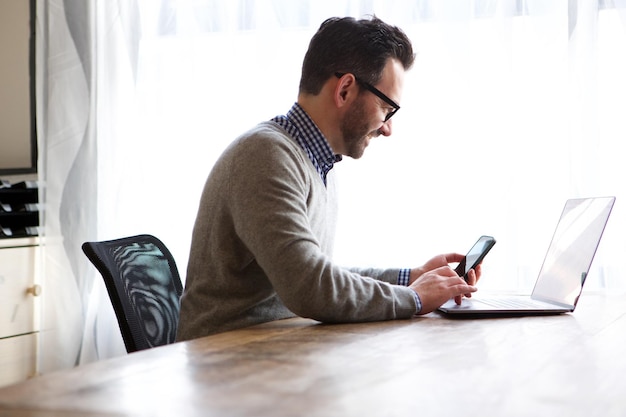 Oudere man die thuis werkt met laptop en mobiele telefoon