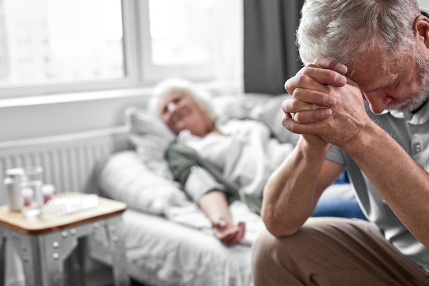 oudere man huilend en rouwend om het verlies van zijn vrouw, zittend aan haar zijde. focus op boos man naar beneden te kijken. coronavirus, covid-19 concept